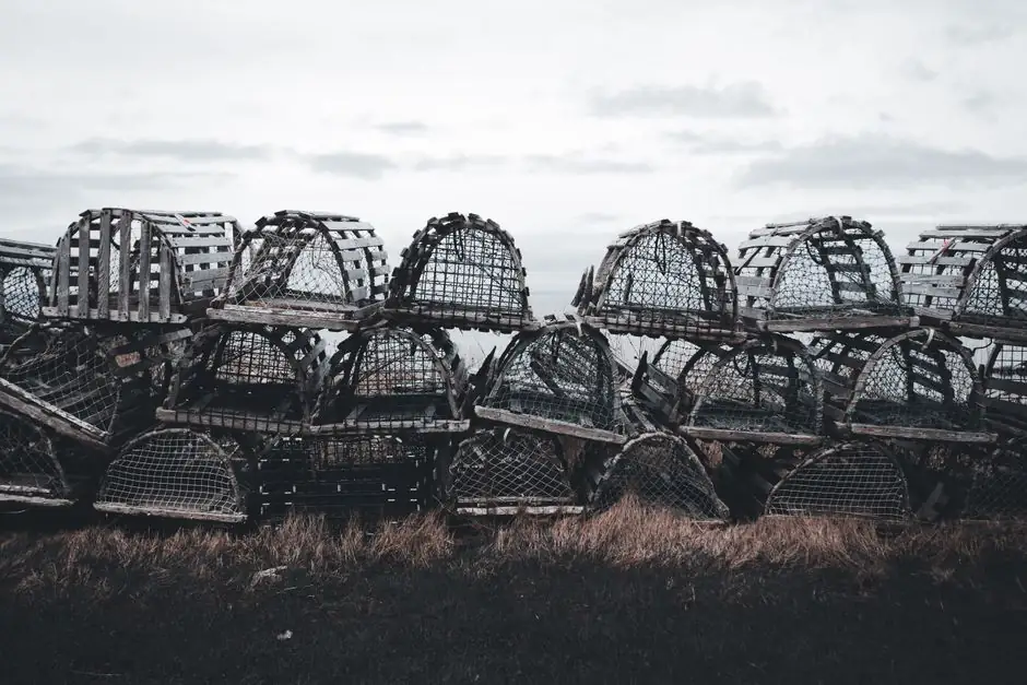 A collection of rustic wooden lobster traps stacked in a coastal field with a cloudy sky.